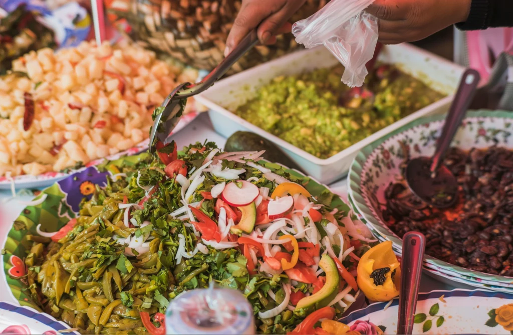 A catering spread laid out on a table: nopales salad with radish and chilli, guacamole, chicharrones and beans, being served with tongs.